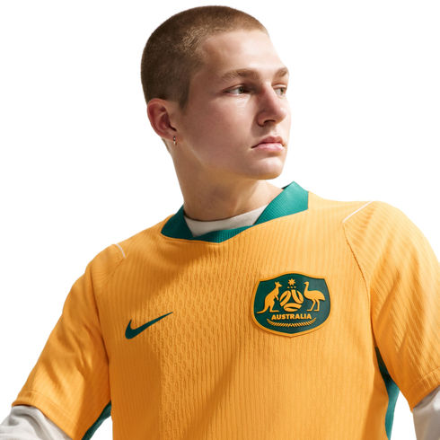 Man with a buzz cut wearing a yellow and green Australian soccer jersey, looking to the side against a solid black background.