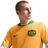 Man with a buzz cut wearing a yellow and green Australian soccer jersey, looking to the side against a solid black background.