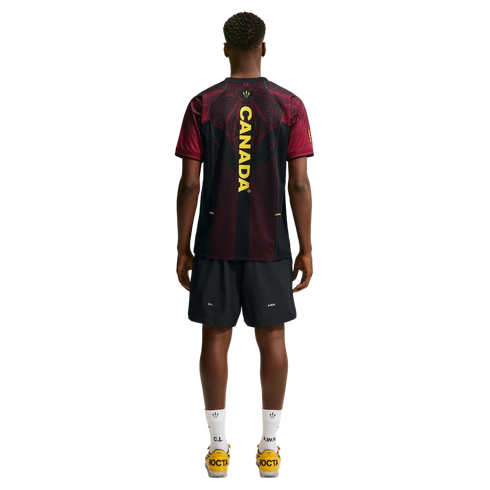 Rear view of a man wearing a black and red jersey with "CANADA" on the back, black shorts, and yellow shoes against a white background.