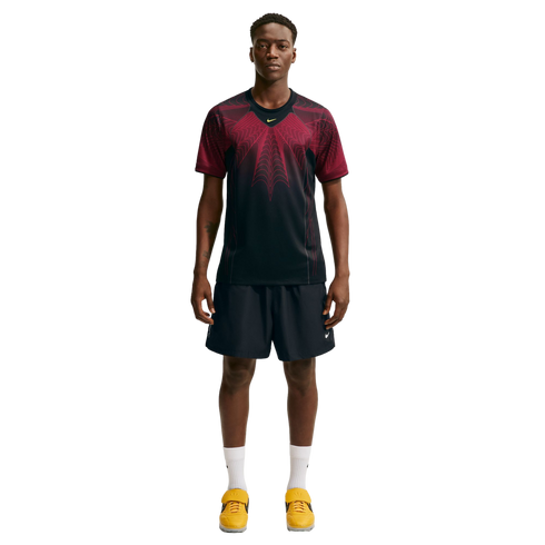 Full-body shot of a man wearing a black and maroon patterned athletic t-shirt, black shorts, white socks, and yellow sneakers, standing against a white background.