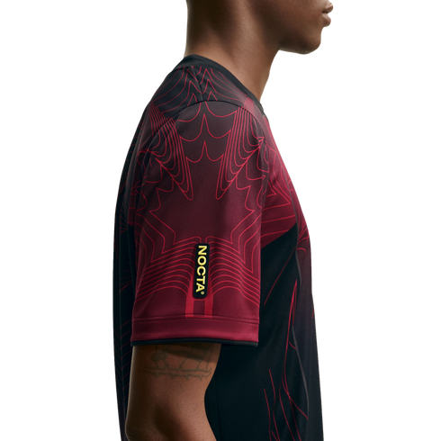 Side profile of a person wearing a maroon and black jersey with graphic line patterns and a yellow "NOCTA" label on the sleeve.