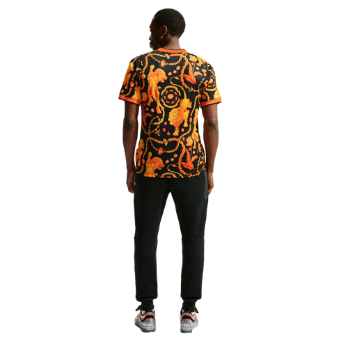 Back view of a man wearing a black t-shirt with a bold orange and yellow lion and chain pattern, black sweatpants, and silver sneakers.