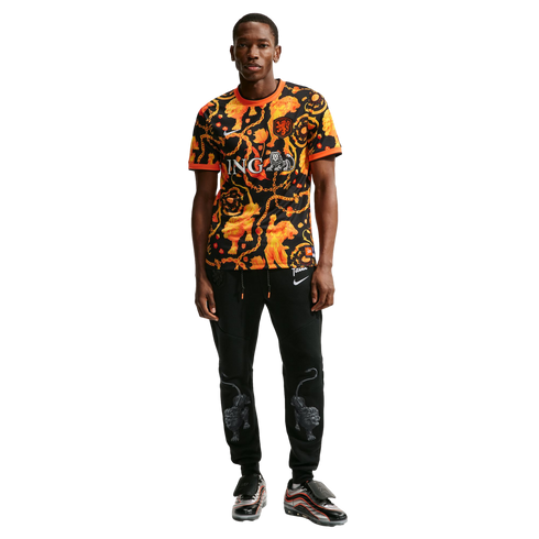 A man poses in a patterned orange and black jersey, black joggers featuring silver lion graphics, and silver Nike sneakers, all against a white background.