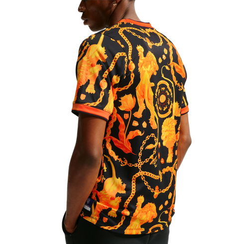 Back view of a person wearing a black t-shirt with an ornate orange pattern of lions, chains, and tulips against a white background.
