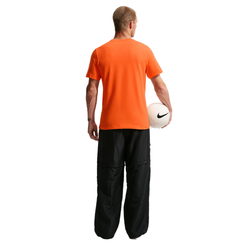 A rear view of a man standing against a black background, wearing a bright orange T-shirt and baggy black pants. He holds a white Nike soccer ball under his left arm.
