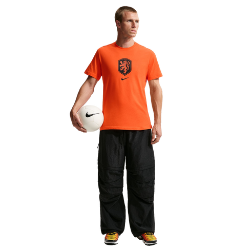 A man stands against a solid black background wearing a bright orange Nike t-shirt featuring the Netherlands national team crest, baggy black textured pants, and colorful sneakers. He holds a white Nike soccer ball under his right arm.