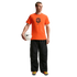 A man stands against a solid black background wearing a bright orange Nike t-shirt featuring the Netherlands national team crest, baggy black textured pants, and colorful sneakers. He holds a white Nike soccer ball under his right arm.