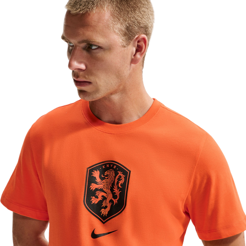 A man with short blond hair wearing a bright orange Nike t-shirt featuring a black KNVB lion crest. He is looking off to the side against a solid black background.