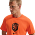 A man with short blond hair wearing a bright orange Nike t-shirt featuring a black KNVB lion crest. He is looking off to the side against a solid black background.