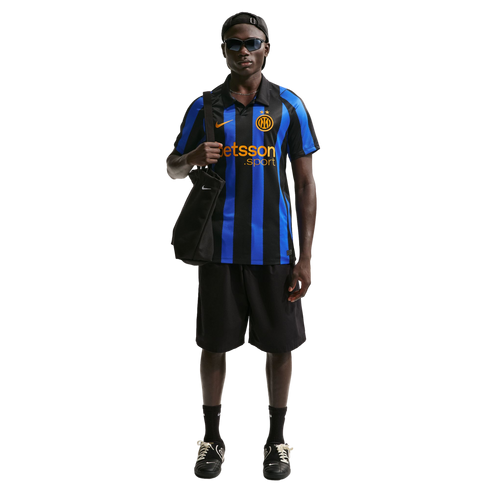 A Black man models an Inter Milan soccer jersey, black shorts, and sunglasses, carrying a black tote bag against a white background.