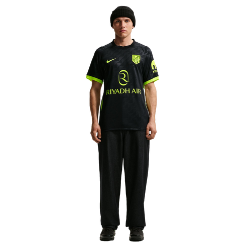 Man in a black Nike Atletico Madrid jersey with neon accents and "RIYADH AIR" sponsor, paired with black pants and a beanie, standing against a white background.