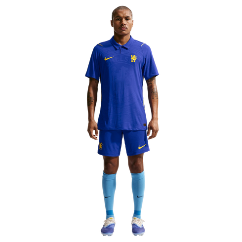 A man stands against a white background wearing a blue Nike Chelsea FC football kit with a polo-style jersey, matching blue shorts, and light blue socks.
