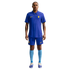A man stands against a white background wearing a blue Nike Chelsea FC football kit with a polo-style jersey, matching blue shorts, and light blue socks.