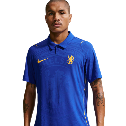 A man wearing a blue, collared Chelsea FC jersey with yellow logos and a large textured crest on the chest, isolated on a white background.