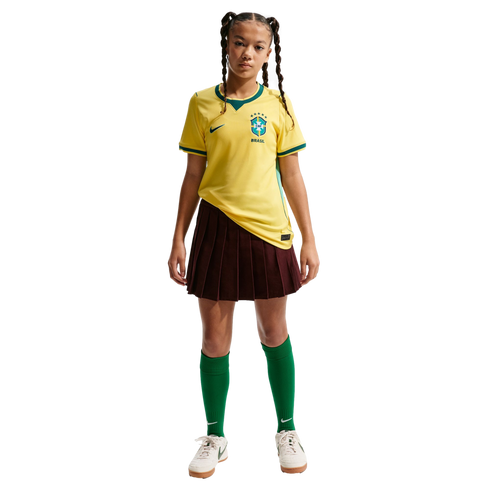 A young girl wearing a yellow Brazil soccer jersey, a maroon pleated skirt, green socks, and white sneakers, standing against a black background.