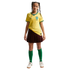 A young girl wearing a yellow Brazil soccer jersey, a maroon pleated skirt, green socks, and white sneakers, standing against a black background.
