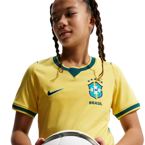 A person with braided hair wearing a yellow Brazil soccer jersey and holding a soccer ball against a black background.