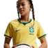 A person with braided hair wearing a yellow Brazil soccer jersey and holding a soccer ball against a black background.