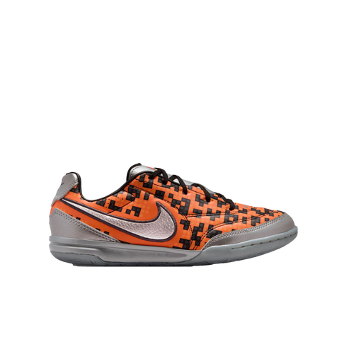 Side profile of an orange and black pixelated Nike indoor soccer shoe with metallic silver accents on a white background.