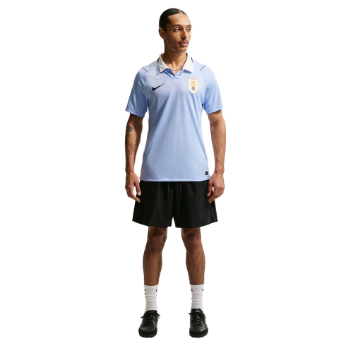 A man wearing a light blue Uruguay soccer jersey, black shorts, and white socks, standing against a black background.