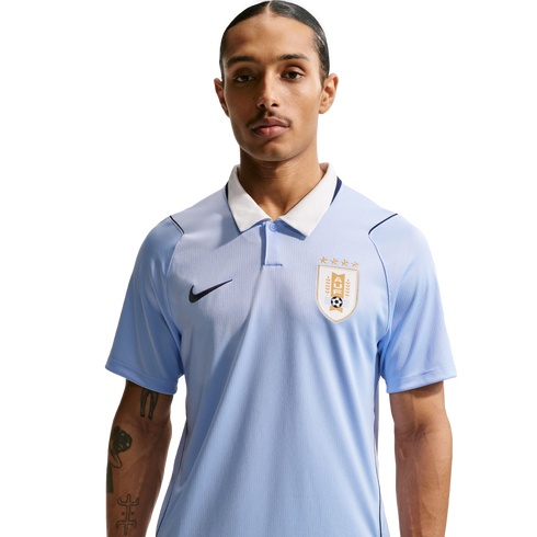 A man with a mustache wears a light blue Uruguay national football team polo shirt with a white collar and Nike logo against a black background.