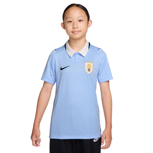 A young girl wears a light blue Nike Uruguay soccer jersey with a white collar and black shorts against a solid black background.