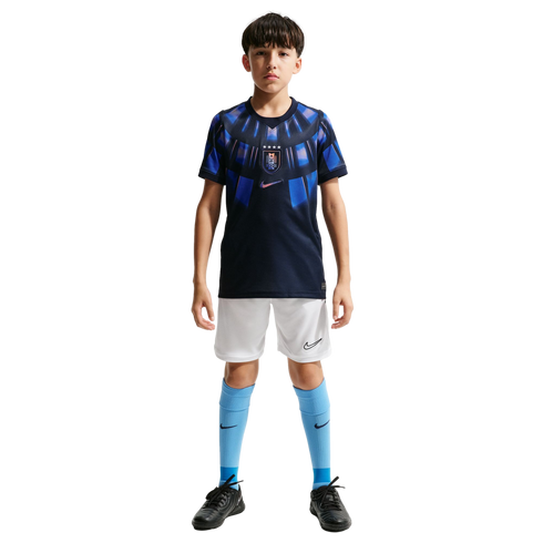 A young boy stands against a solid black background wearing a navy and light blue Nike soccer kit, white shorts, light blue socks, and black soccer cleats.