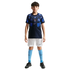 A young boy stands against a solid black background wearing a navy and light blue Nike soccer kit, white shorts, light blue socks, and black soccer cleats.