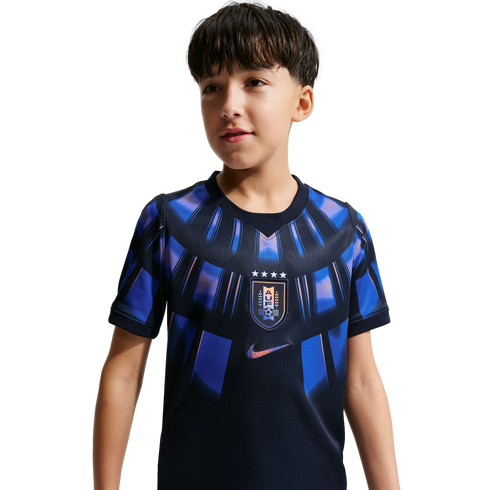 A young boy wearing a navy blue Uruguay national team soccer jersey with iridescent blue and purple patterns. The jersey features the AUF crest on the chest and a Nike swoosh logo, set against a solid black background.