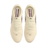 A top-down view of a pair of off-white Nike soccer cleats against a solid black background. The cleats feature cream-colored uppers and laces, with dark reddish-brown insoles containing white text and logos.