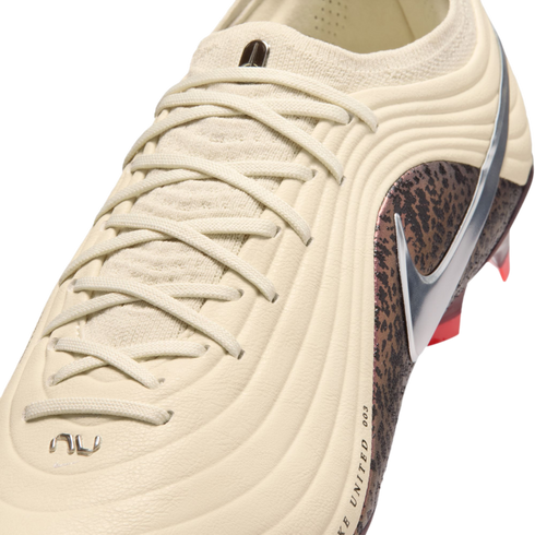 A close-up shot of a cream-colored Nike soccer cleat with a textured ribbed upper and matching laces. A metallic silver swoosh sits atop a dark brown mottled pattern on the side. The background is solid black.