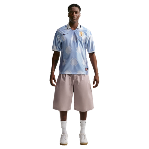 A man wearing a light blue sun-patterned soccer jersey, tan shorts, and white sneakers stands against a plain white background.