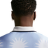 Back view of a person with short curly hair and gold hoop earrings, wearing a light blue polo shirt with a white sun pattern.
