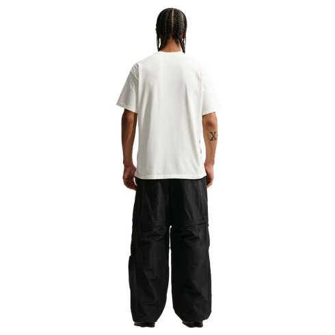 A full-body rear view of a man with braided hair standing against a solid black background. He is wearing a plain white short-sleeved t-shirt and oversized, black technical pants. A small "X" tattoo is visible on his right forearm.