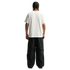 A full-body rear view of a man with braided hair standing against a solid black background. He is wearing a plain white short-sleeved t-shirt and oversized, black technical pants. A small "X" tattoo is visible on his right forearm.