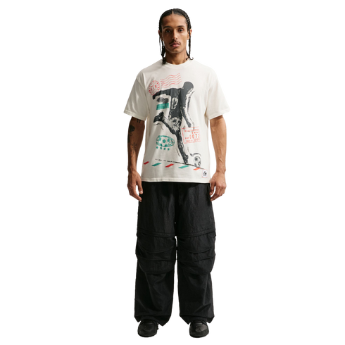 A man stands against a solid black background wearing a white graphic t-shirt featuring a soccer player silhouette, oversized black cargo pants, and chunky black sneakers.
