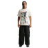 A man stands against a solid black background wearing a white graphic t-shirt featuring a soccer player silhouette, oversized black cargo pants, and chunky black sneakers.