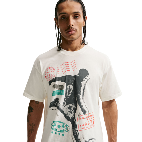 A young man with braided hair and a thin mustache wearing a cream-colored T-shirt with a large soccer-themed graphic, set against a solid black background.