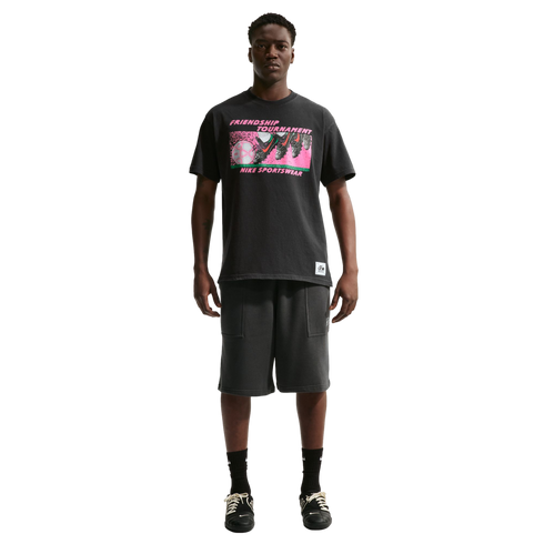 A man stands against a white background wearing a black Nike "Friendship Tournament" graphic t-shirt, charcoal shorts, and black sneakers.