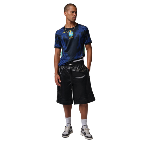 A male model wears a blue Brazil national team soccer jersey, black shiny shorts, and Jordan sneakers, standing against a black background.
