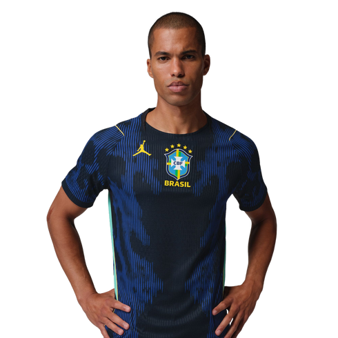 Man in a navy blue Brazil soccer jersey featuring a yellow Jordan logo and team crest, standing with hands on hips against a black background.