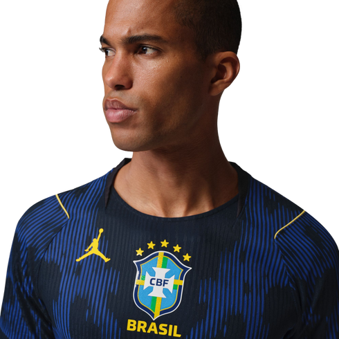 A man in a blue Brazil soccer jersey with yellow Jordan and CBF logos, looking sideways against a black background.