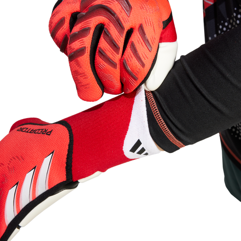 Close-up of a person putting on red Adidas Predator goalkeeper gloves, showing the textured grip and white brand logo.