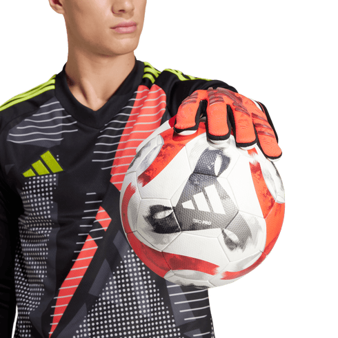 Goalkeeper in a black, red, and yellow jersey holding an Adidas Tiro Pro soccer ball with an orange-gloved hand against a green background.