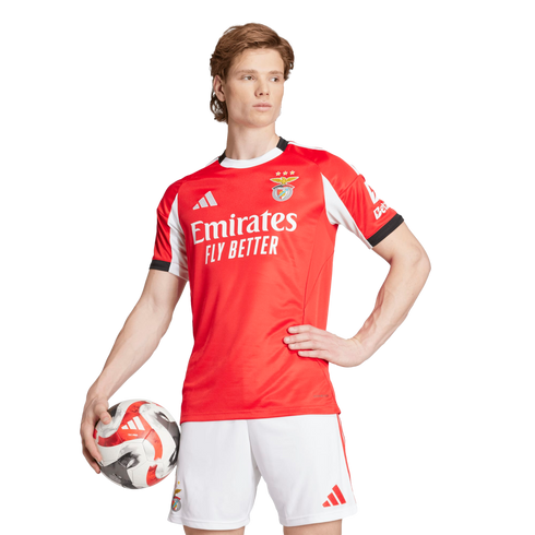 A male athlete posing in a red SL Benfica home jersey and white shorts while holding a soccer ball.