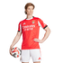 A male athlete posing in a red SL Benfica home jersey and white shorts while holding a soccer ball.