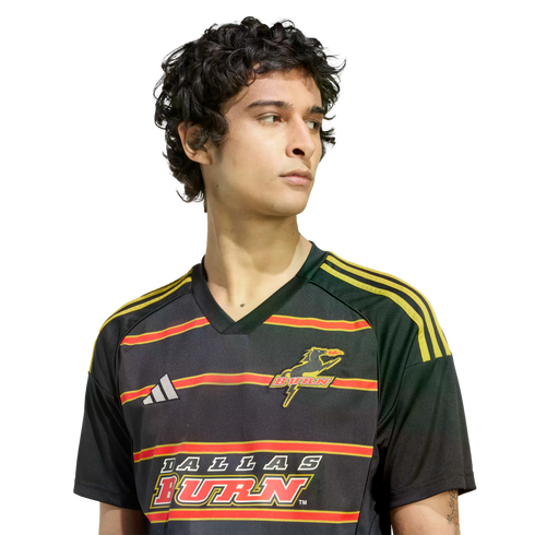 A young man with dark curly hair wearing a black Adidas Dallas Burn soccer jersey with red and yellow horizontal stripes, looking to the side.