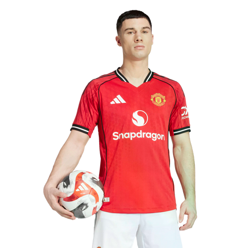 A man in a red Manchester United home jersey with the Snapdragon logo holds a soccer ball against a solid black background.