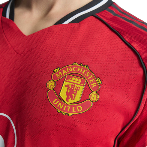 Close-up of a red Manchester United football jersey featuring the team's yellow and red crest on the chest, with black shoulder stripes and patterned fabric.