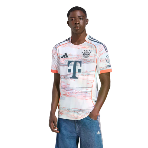 Young Black man modeling a white FC Bayern Munich jersey with a red and grey abstract pattern and blue denim shorts against a white background.